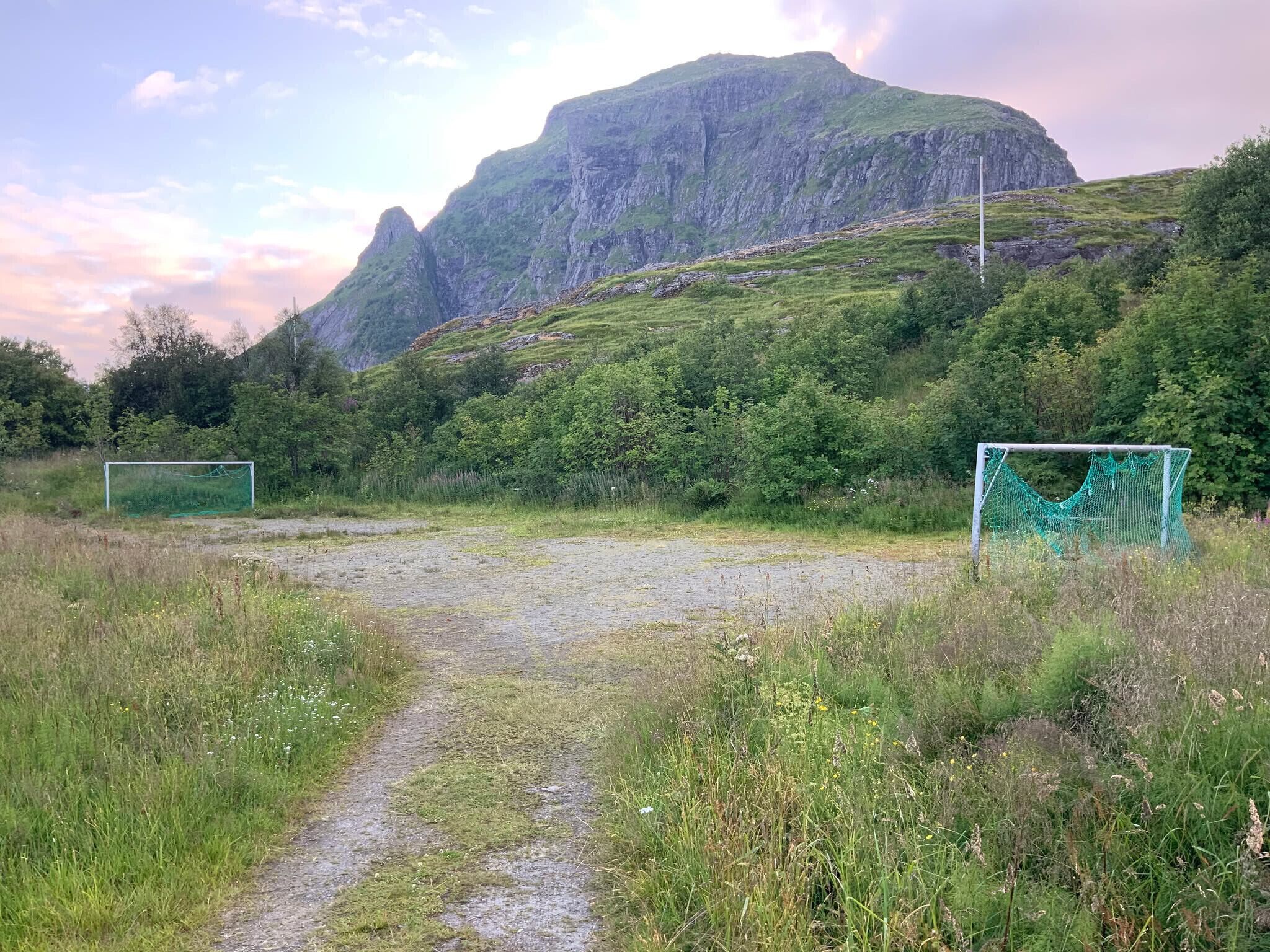 Een landelijk landschap met een onverhard pad dat leidt naar een voetbaldoel, omringd door groen en een berg op de achtergrond.
