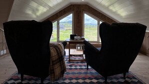 Ein gemütliches Dachzimmer mit geneigter Decke, zwei Sesseln, einem kleinen Tisch mit Lampe und Blick auf eine grüne Landschaft durch große Fenster.