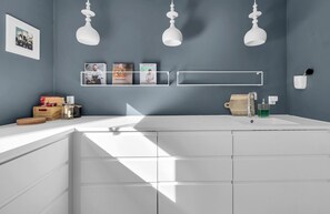 Um banheiro moderno com toucador branco, pia e espelho. Há três luminárias pendentes, um quadro na parede e uma prateleira com livros e porta-escovas de dentes.