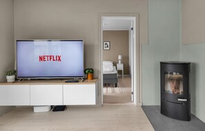 Ett modernt vardagsrum med en platt-TV som visar "NETFLIX", ett vitt skåp, en svart vedkamin och en dörröppning som leder till ett sovrum.