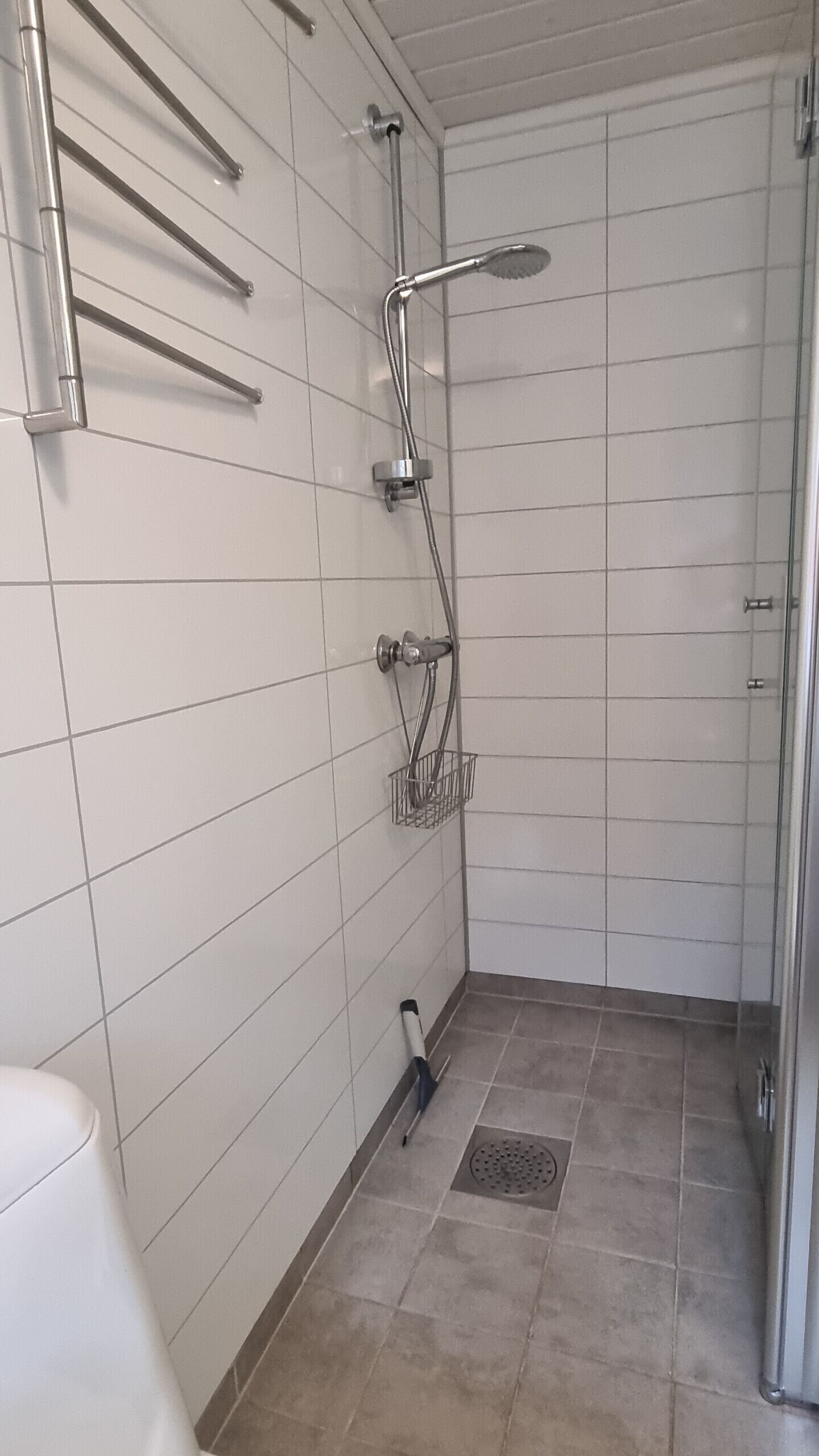 A bathroom with white tiled walls, a shower with a handheld showerhead, a handheld showerhead on the floor, and a metal towel rack.
