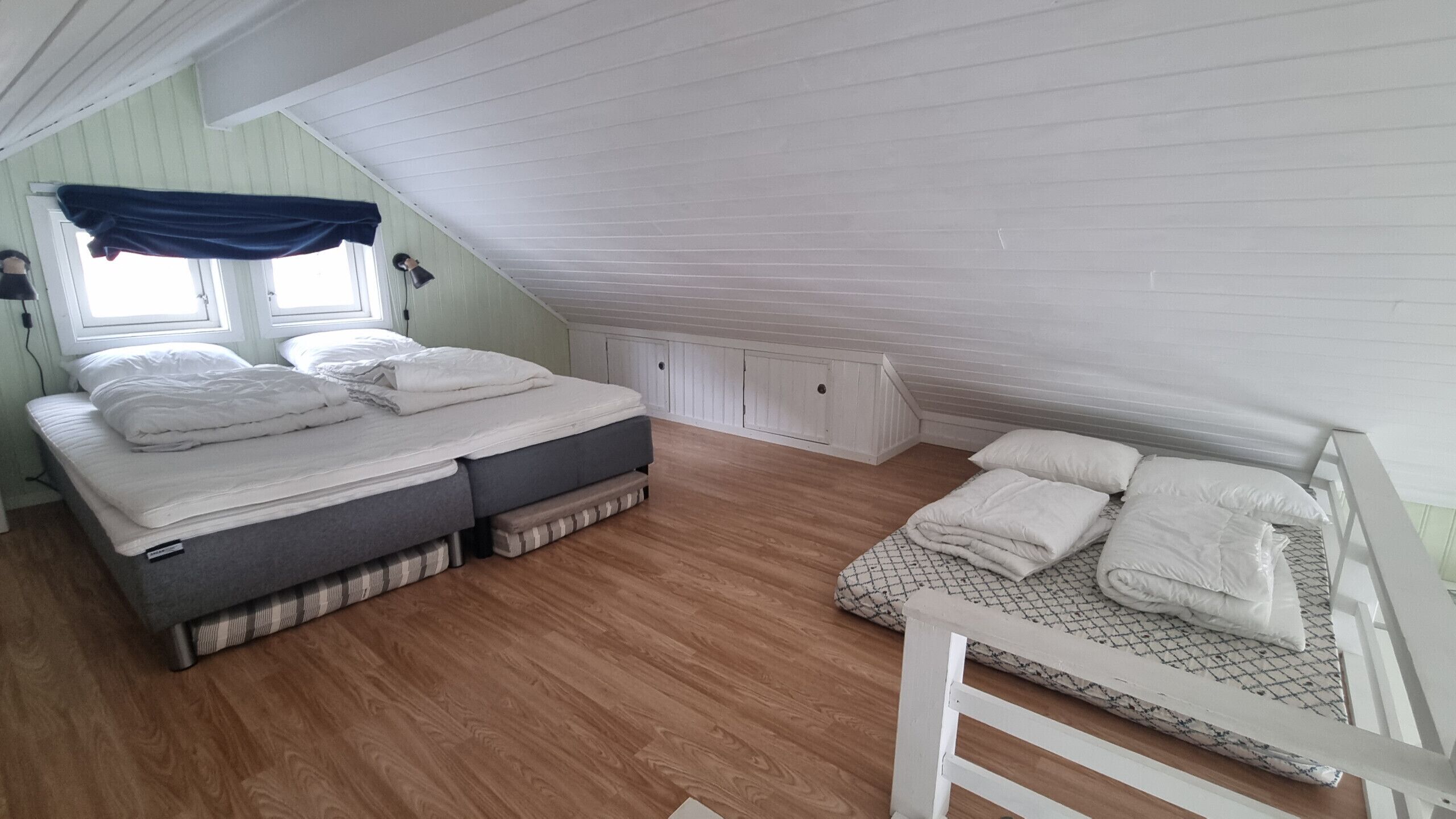Une chambre située dans un espace de type « loft », avec un plafond en pente, un lit avec un matelas et des oreillers, un banc avec des coussins et une fenêtre avec des rideaux.