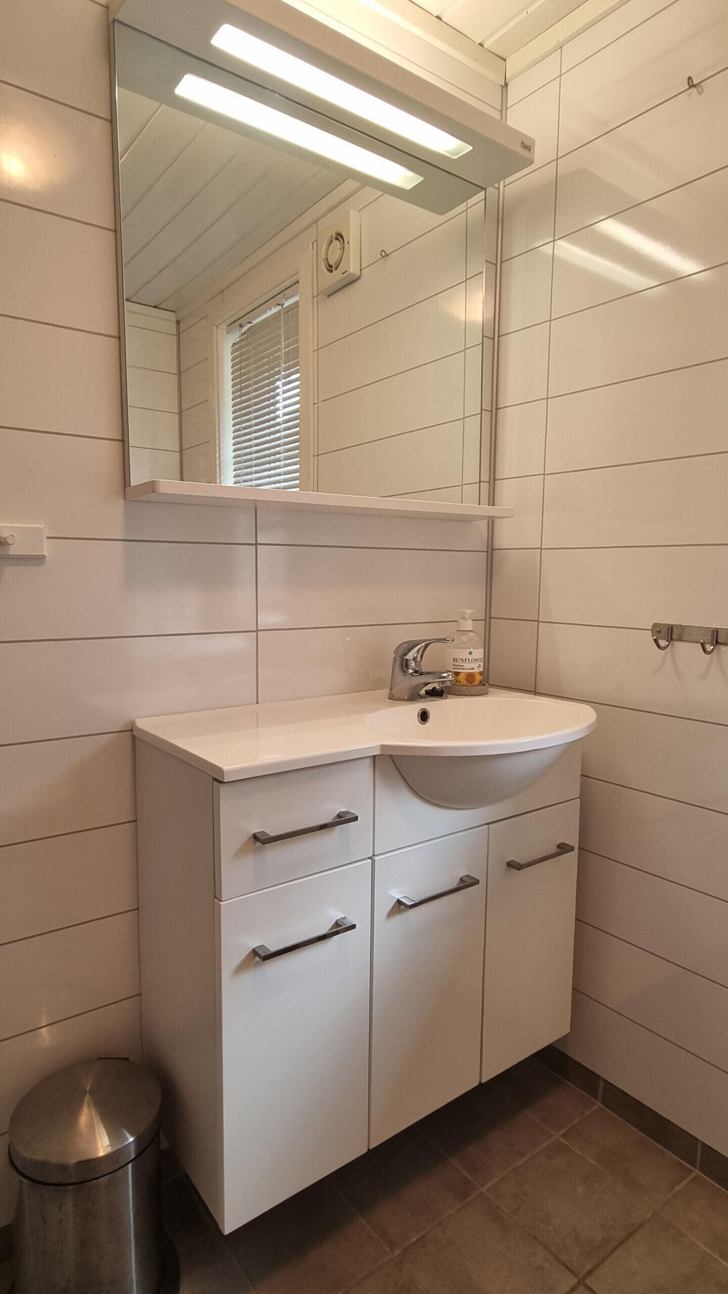 A bathroom with a white sink, mirror, and cabinet, a trash bin, and a window with blinds.