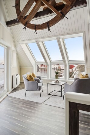 Una habitación con techos altos, tragaluces, un radiador blanco, sillas grises, una pequeña mesa y vistas a un paisaje nevado.