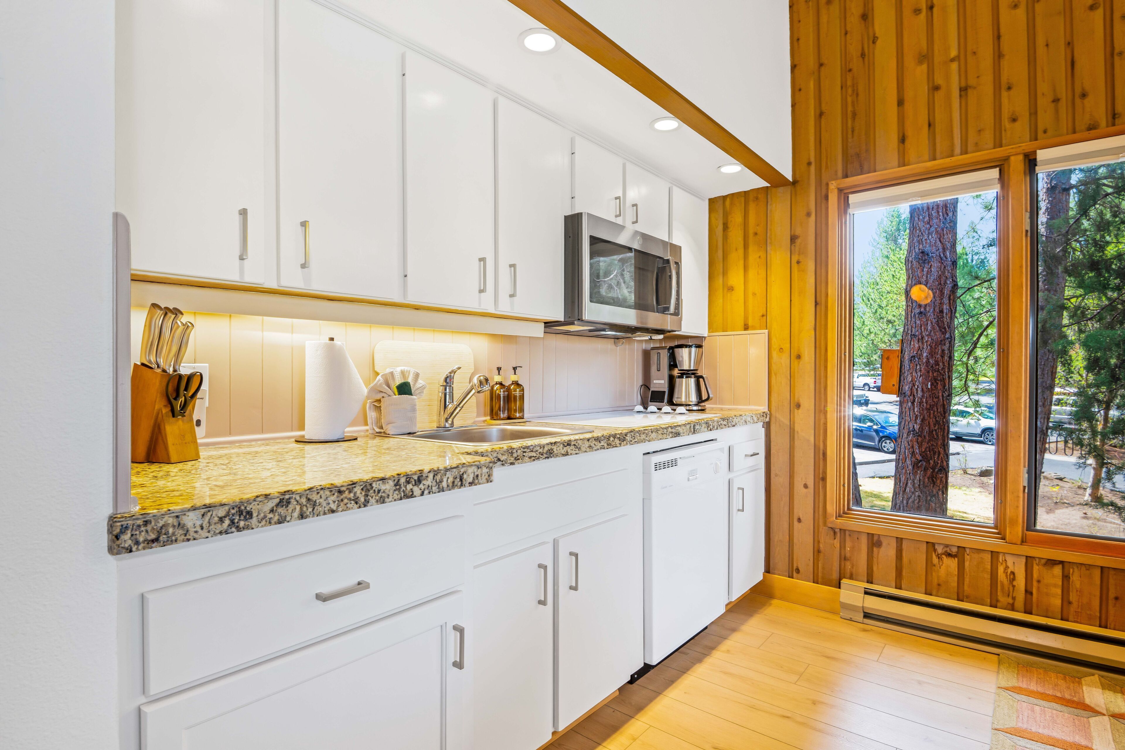 A modern kitchen with white cabinets, a granite countertop, a built-in microwave, and a window with a view of trees.