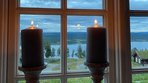 Une fenêtre avec deux bougies posées sur des chandeliers, une vue sur un lac et une maison en arrière-plan.