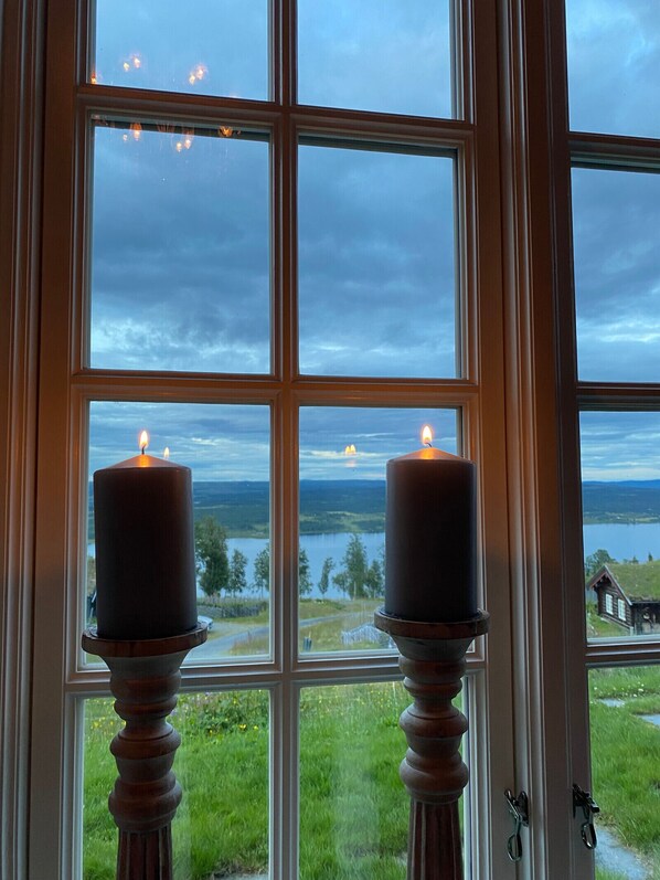 Ein Fenster mit zwei Kerzen auf einem Sockel, Blick auf einen See und ein Haus im Hintergrund.