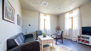 A living room with a sofa, chairs, a coffee table, a TV stand with books, and two windows with curtains.
