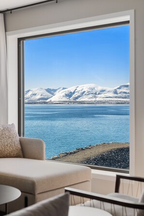 Ein Zimmer mit großem Fenster, Blick auf ein Gewässer und schneebedeckte Berge.
