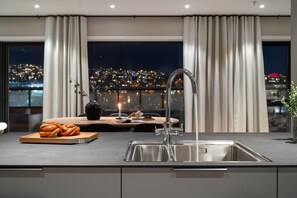 Eine moderne Küche mit Spüle aus Edelstahl, einem Holztisch mit Gebäck und Blick auf die Stadt durch große Fenster.