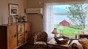 A room with a wooden cabinet, a floral armchair, a table with a lamp, and a view of a red barn and greenery.