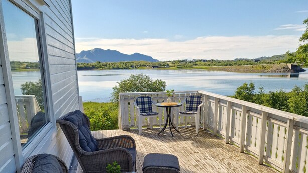 A deck with wicker furniture, a small table, and chairs overlooking a lake with mountains in the background.