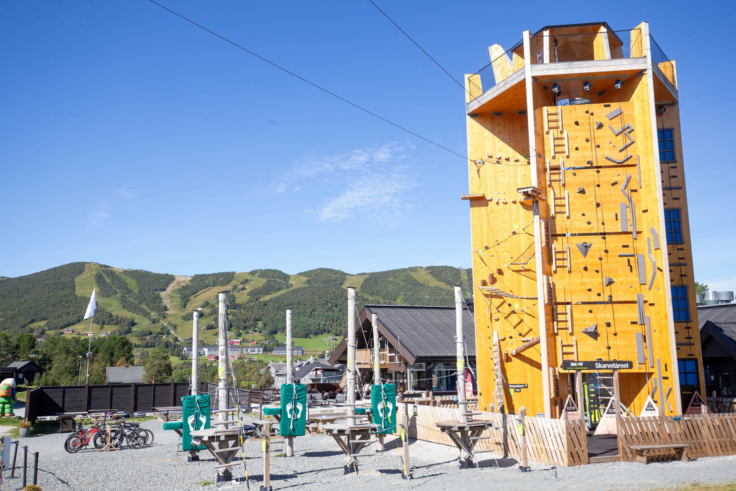 A climbing wall facility with a wooden climbing wall, a sign reading 'Skarvetätmet,' and a view of a mountainous landscape in the background.