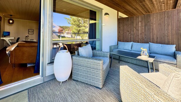 Un área de estar exterior moderna con mobiliario de mimbre, un ventanal y vistas del paisaje circundante.