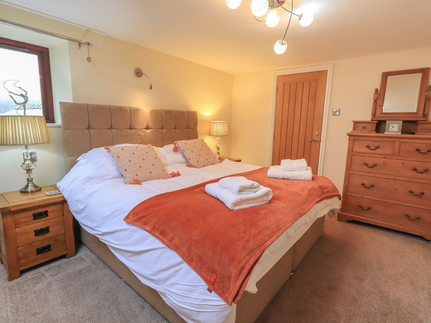 A neatly made bed with pillows, a bedside lamp, a wooden dresser, and a door.