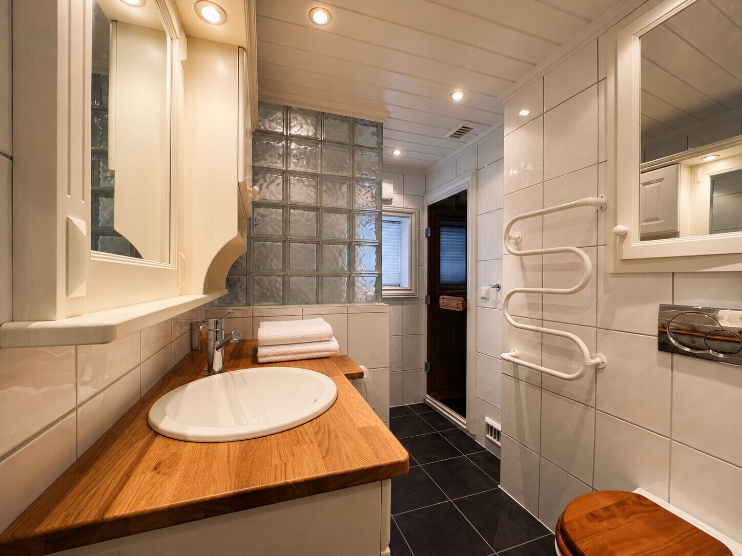 Casa de banho moderna com toucador de madeira, cabine de duche em vidro e chão em azulejo branco.