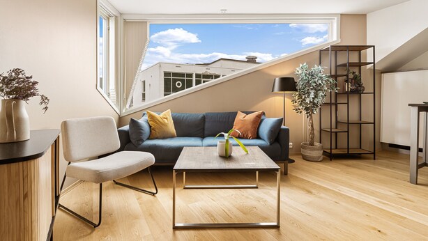 Una sala de estar moderna con un sofá, una mesa de centro y una silla. Hay un ventanal que ofrece vistas a edificios y un cielo despejado.