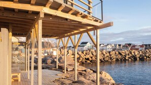 A wooden deck with a metal railing, overlooking a rocky shoreline and residential buildings.