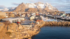 A coastal town with buildings along the waterfront, a rocky shoreline, and snow-capped mountains in the background.