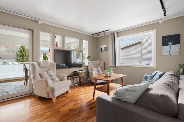 A living room with a sofa, armchair, and a television. There is a wooden table and a window with blinds. - Trivelig leilighet i rolige, bynære omgivelser (Tromsø)