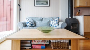A room with a wooden table, a sofa with patterned cushions, a framed picture on the wall, and a wood-burning stove.