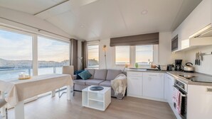 A modern interior with a dining area, sofa, kitchen, and large windows overlooking a body of water.