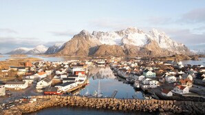 A coastal town with buildings along the waterfront, a harbor with boats, and snow-capped mountains in the background.