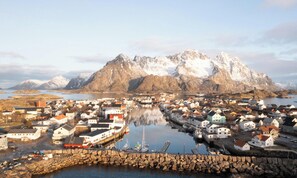Un pueblo costero con edificios a lo largo del paseo marítimo, un puerto con barcos y montañas nevadas al fondo.