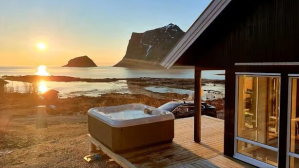 A wooden cabin with a hot tub and a view of the ocean and mountains at sunset.