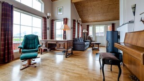 A spacious living area with a wooden ceiling, large windows, a teal armchair, a sofa, a piano, and a heater.