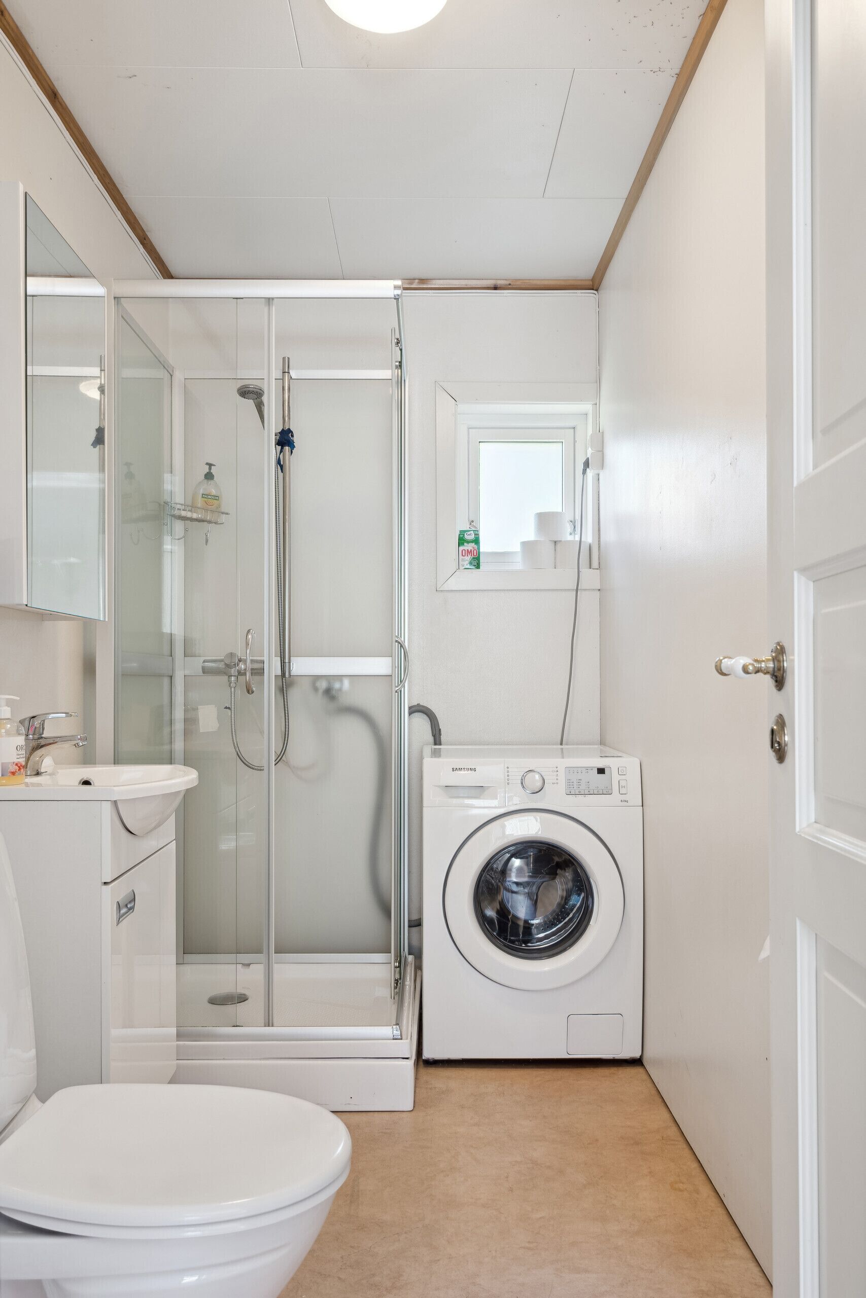 A compact bathroom with a glass-enclosed shower, a washing machine, a toilet, and a mirror.