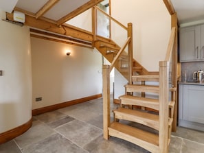 Eine Holztreppe mit Glasgeländern führt zu einer zweiten Etage, einem Küchenbereich mit Schränken und einem Wasserkocher sowie einem gefliesten Boden.