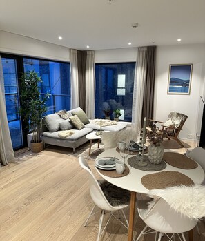 A modern living room with a sofa, dining table, and chairs, large windows with curtains, and a view of the outdoors. - Modern apartment in Tromsø (Tromsø)