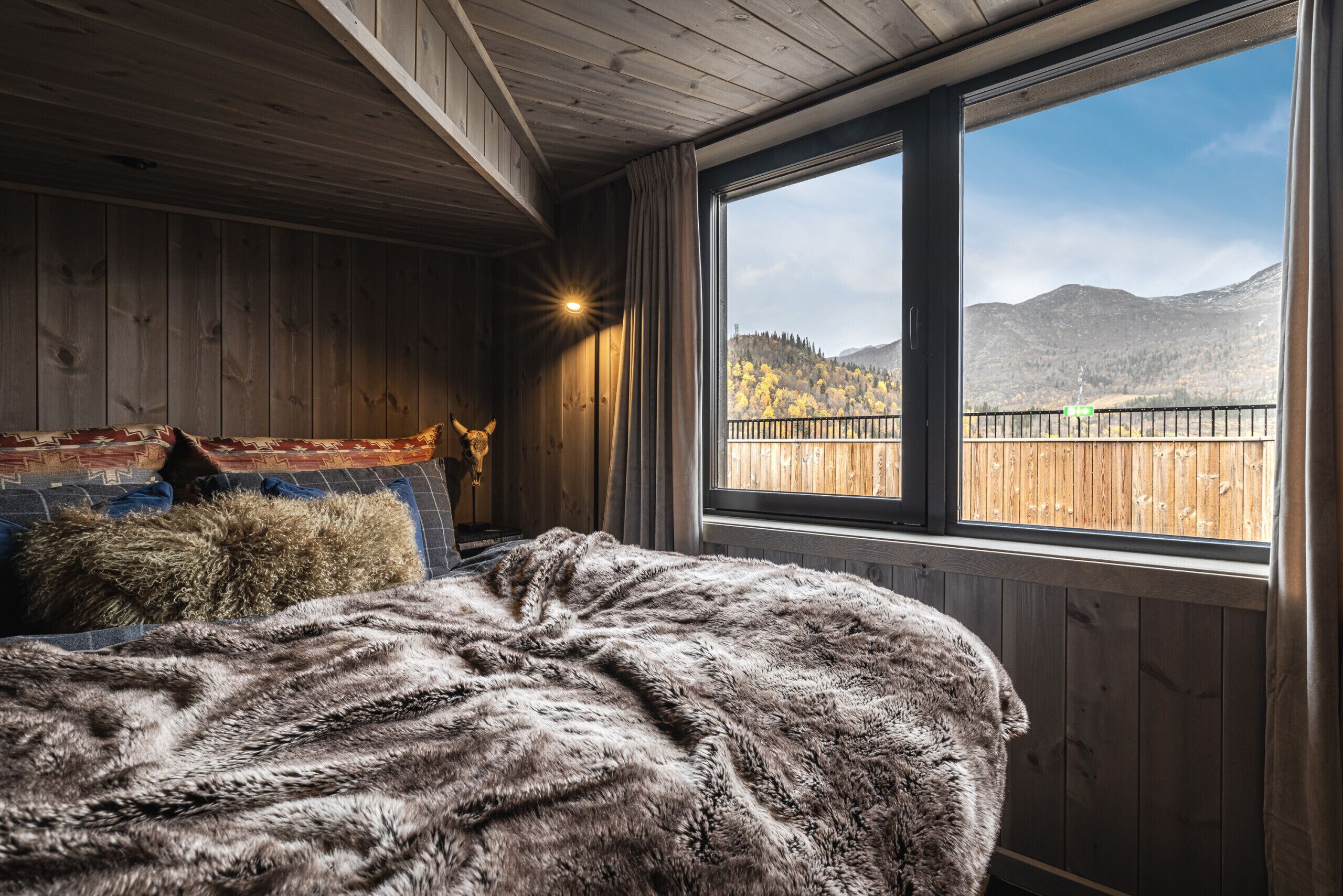 Una habitación con paredes de madera, un ventanal con vistas a montañas, una cama con manta de piel y una lámpara.