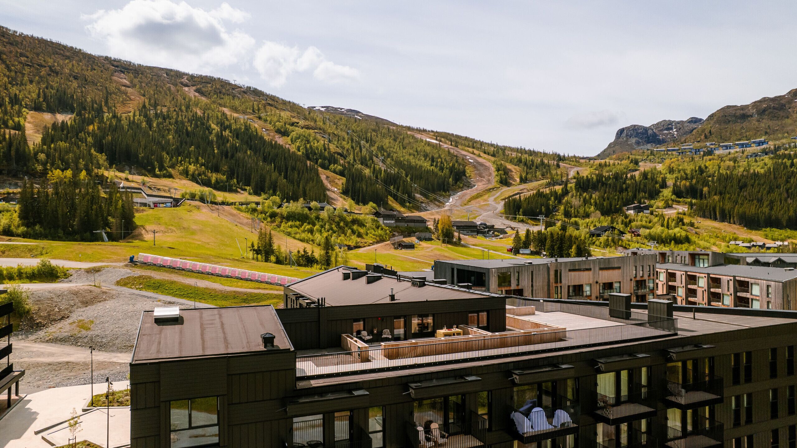 A modern building with a rooftop terrace, a train track, and a mountainous landscape in the background.