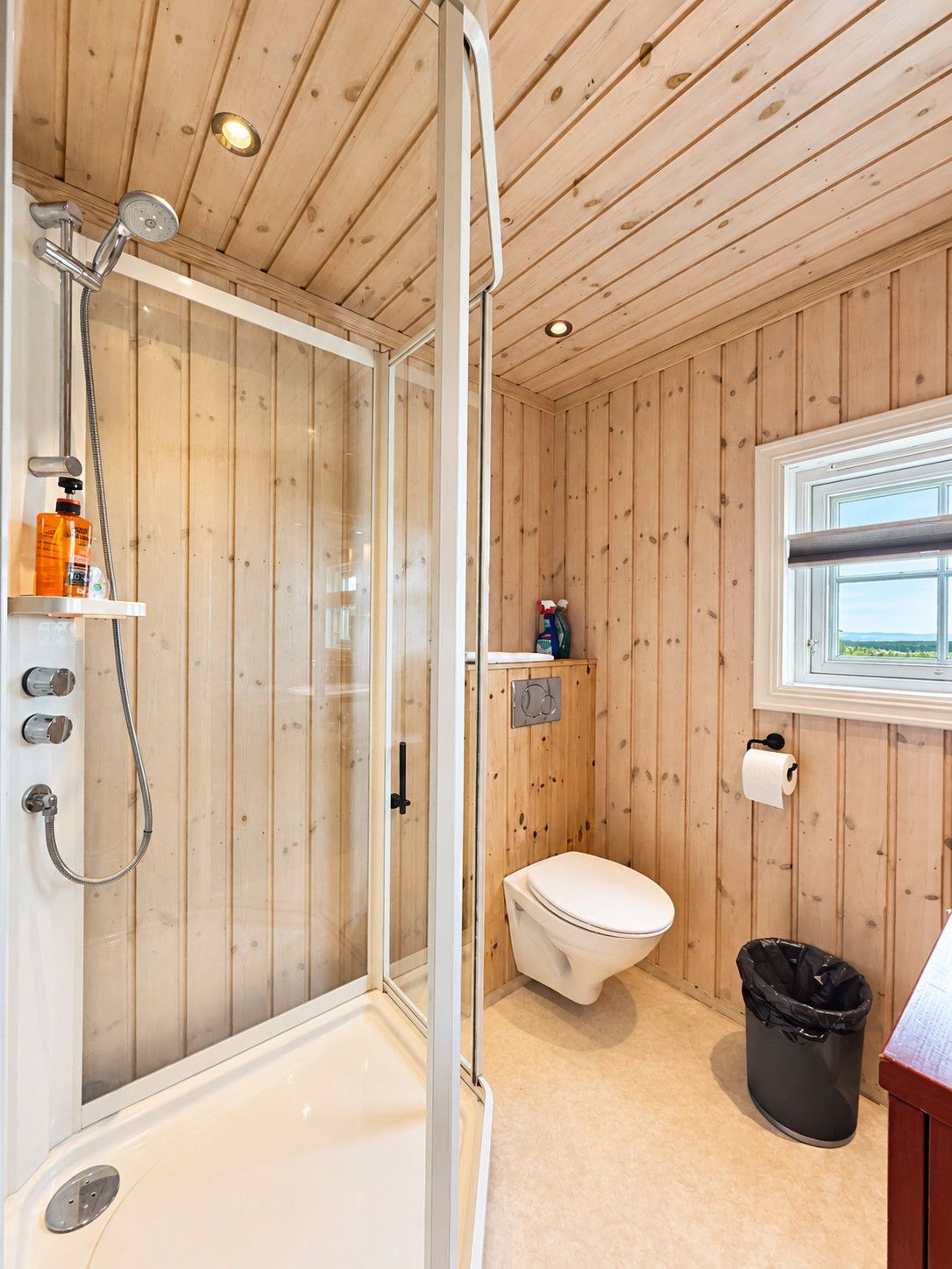 Une salle de bain aux murs lambrissés, dotée d’une douche à parois de verre, de toilettes et d’une fenêtre.