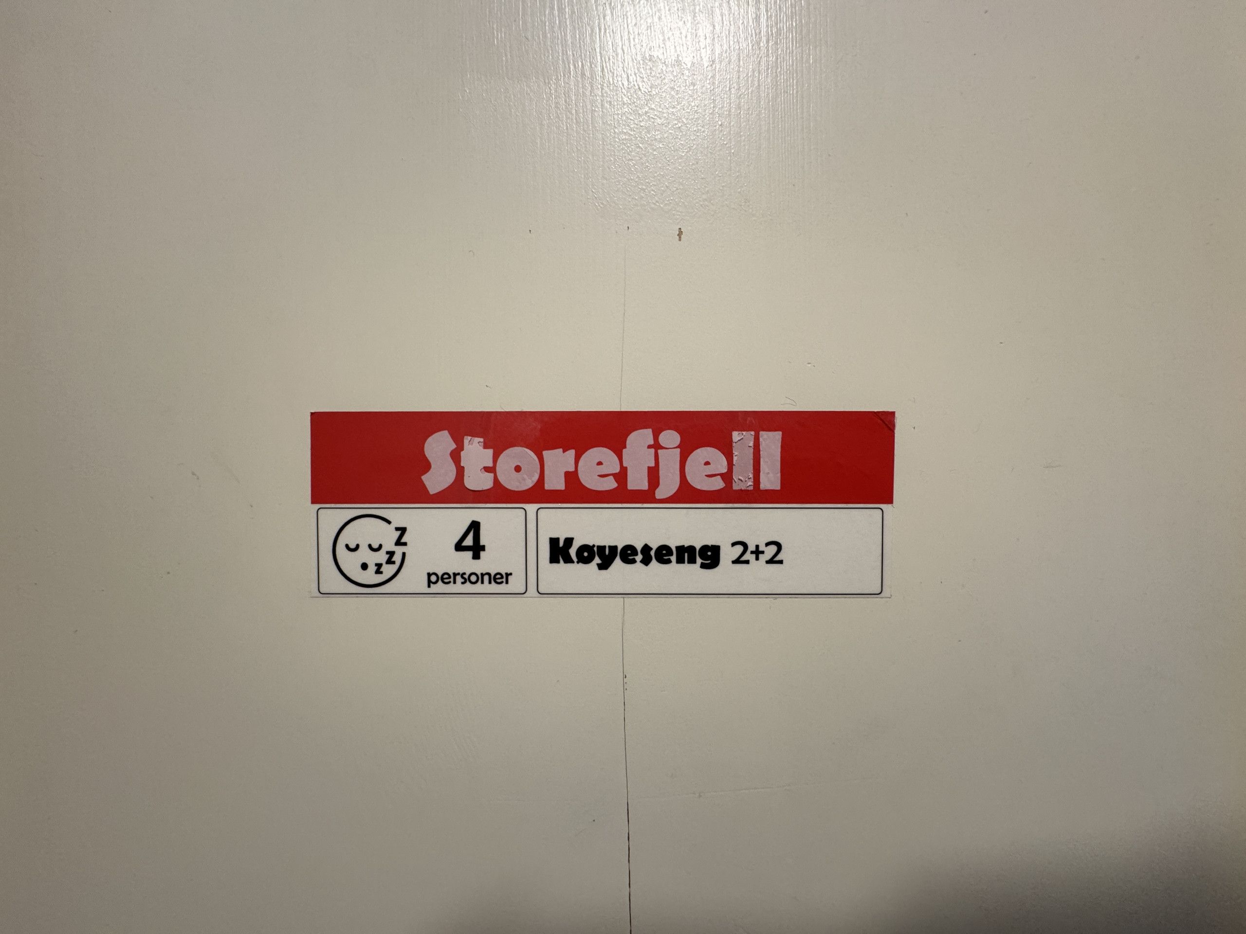 Un autocollant sur un mur portant l’inscription « Storefjell » et « 4 personnes », indiquant le nom du magasin et sa capacité d’accueil.