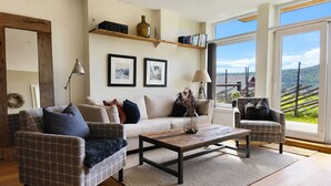 Ein Wohnzimmer mit einer Couch, zwei Sesseln, einem Couchtisch und Blick auf eine ländliche Landschaft durch große Fenster.