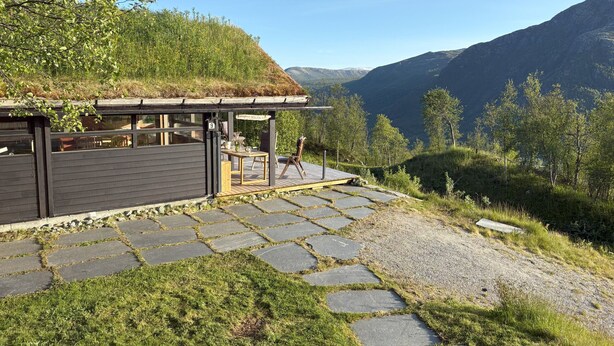A modern building with a stone pathway leading to a covered seating area, surrounded by a mountainous landscape.