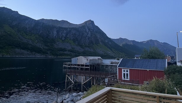 Un muelle de madera se adentra en un lago tranquilo, rodeado de montañas y un edificio rojo.