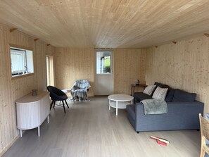 A room with wooden walls and ceiling, a grey sofa, a round white table, a black chair, and a small table with a kettle. - Fantastisk hus med sjøtomt i Gryllefjord (Senja)