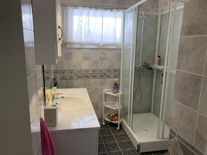 Ein Badezimmer mit einem weißen Waschbecken, einer Duschkabine aus Glas und einem Fenster mit durchsichtigen Vorhängen.