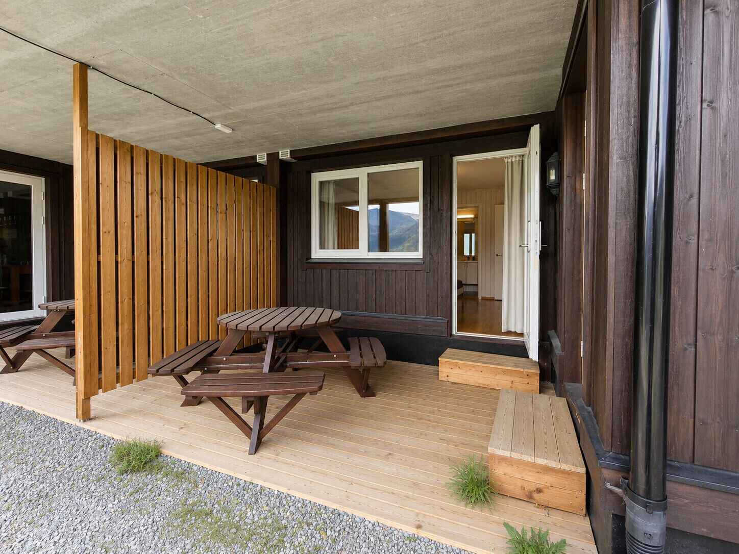 Una terraza de madera con mesas de picnic, una zona cubierta y vistas del interior.