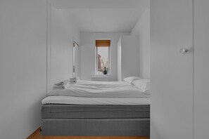 Una habitació estreta amb un llit, una finestra i una porta.