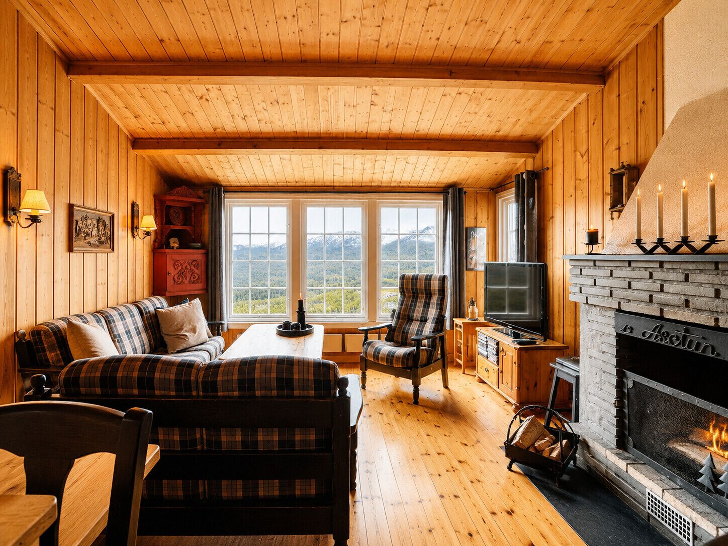 Een gezellig cabin interieur met een houten plafond, ruitjesmeubilair, een open haard, een televisie en een groot raam met uitzicht op de bergen.