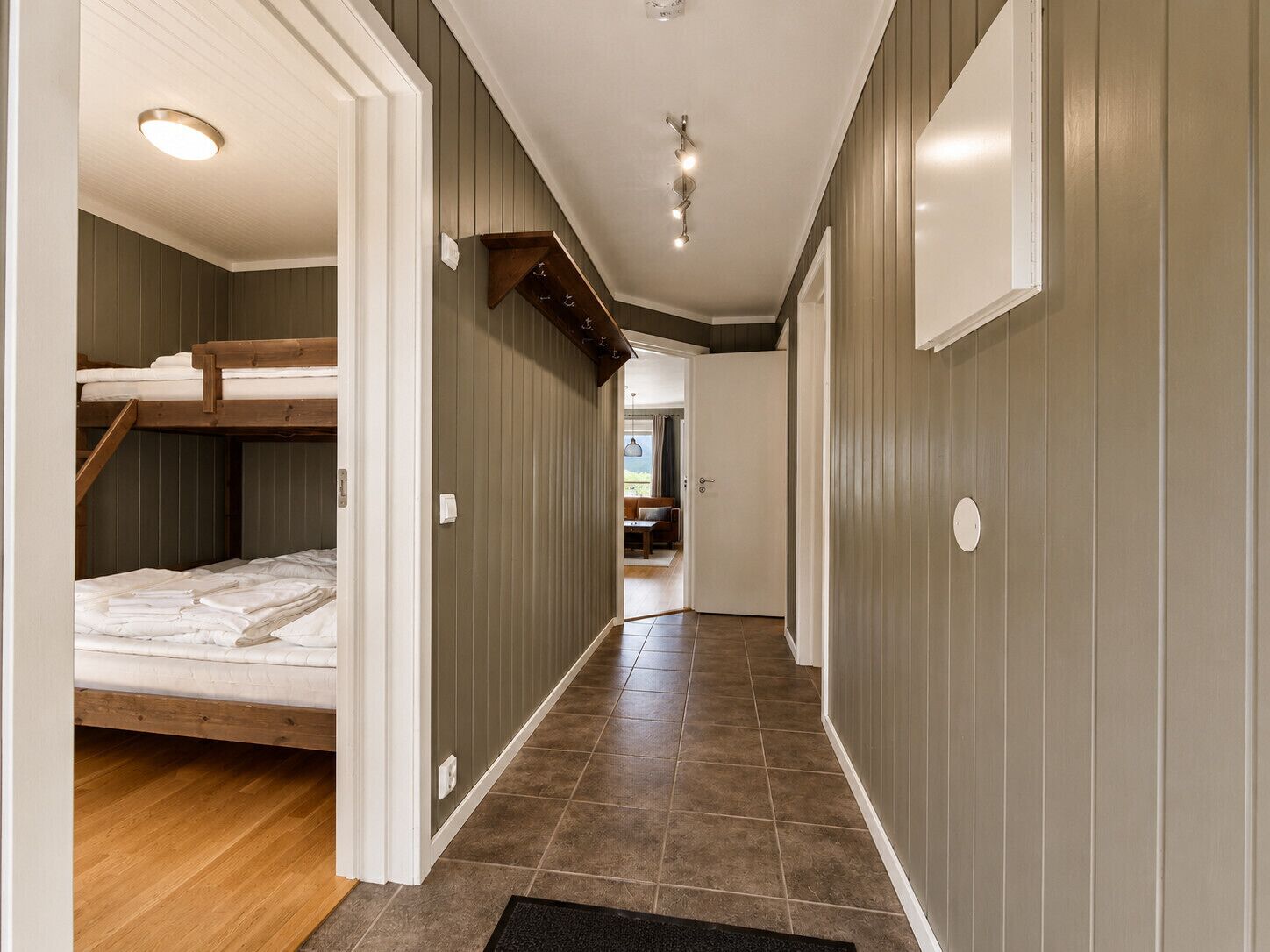 A narrow hallway with a bunk bed, wooden flooring, and a tiled floor leading to a room with a window.