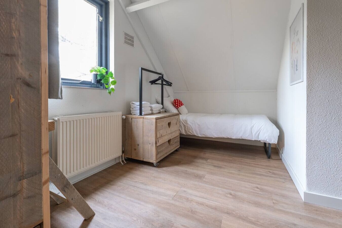Una habitación pequeña y ordenada con una cama, un aparador, un radiador y una ventana con una planta.