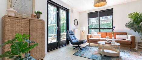 A modern living room with a black leather chair, a brown sofa, and a round coffee table. There is a wicker cabinet and a large window with a view of greenery.