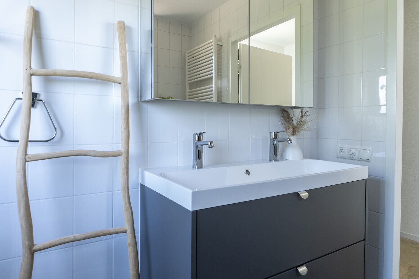 Un baño con un lavamanos blanco, muebles oscuros, un espejo y una escalera de madera.