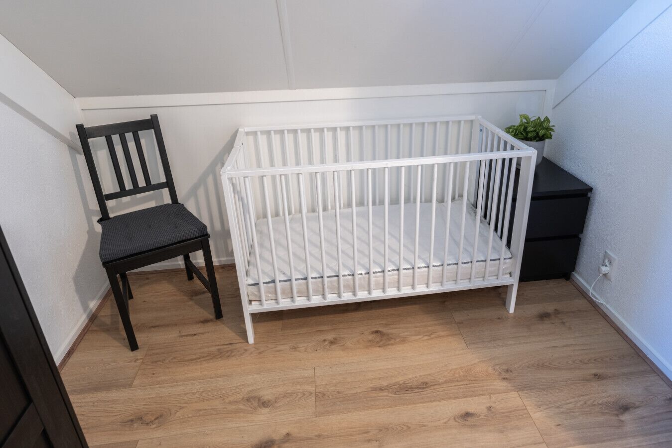Ein weißes Babybett, ein dunkler Holzsessel und eine kleine Pflanze in einer Ecke eines Raumes mit Holzdielen und weißen Wänden.